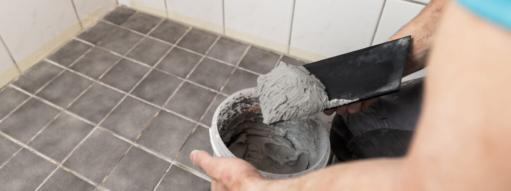 Kylpyhuoneen lattia ja saumauslaastiämpäri, josta asentaja nostaa laastia levittimellä